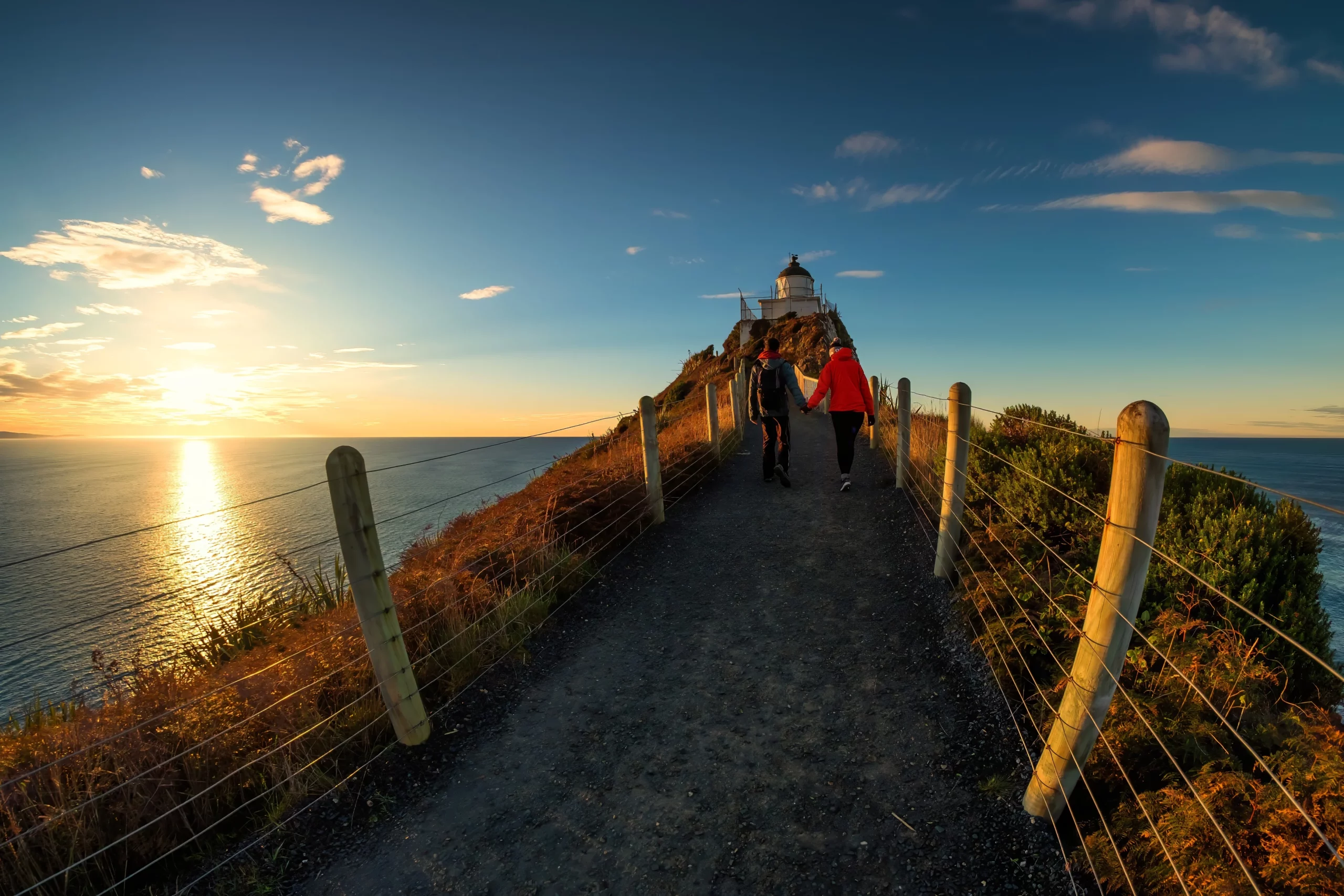 A romantic couple walking toward the lighthouse at Nugget Point, Dunedin, one of the top wedding and honeymoon destinations in New Zealand.